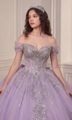 A woman stands in an elegant room with cream-colored walls, wearing the Quinceanera Dress 15730 By Ladivine—a sparkly lavender off-the-shoulder gown featuring a crystal-embellished corset bodice and a glitter-tulle ballgown skirt—her tiara perfectly completing the enchanting look.