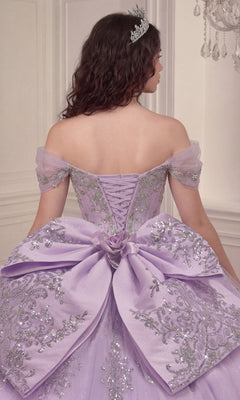 A woman stands facing away in a softly lit, elegant room, wearing the Quinceanera Dress 15730 by Ladivine—a lavender off-the-shoulder gown adorned with silver embroidery and a statement bow. The tiara atop her head complements the crystal-embellished corset bodice and flowing glitter-tulle ballgown skirt, creating an enchanting scene.