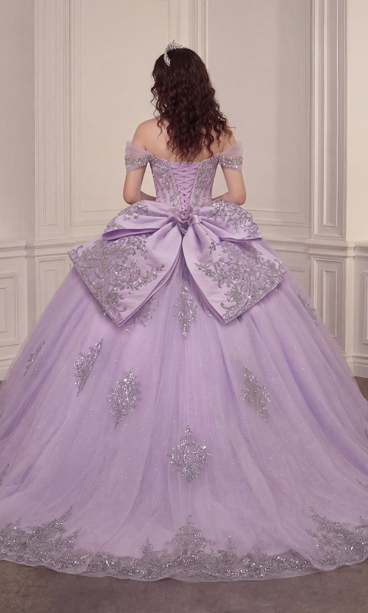 A woman with curly hair stands in an ornate room facing away from the camera, wearing the Quinceanera Dress 15730 By Ladivine—a lavender off-the-shoulder gown adorned with silver embroidery and a large bow, featuring a glitter-tulle ballgown skirt.