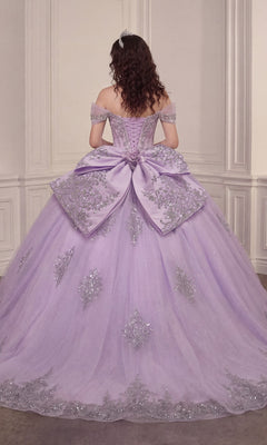 A woman with curly hair stands in an ornate room facing away from the camera, wearing the Quinceanera Dress 15730 By Ladivine—a lavender off-the-shoulder gown adorned with silver embroidery and a large bow, featuring a glitter-tulle ballgown skirt.