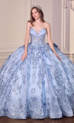 A woman stands indoors against a white wall wearing the Quinceanera Dress 15732 By Ladivine, a strapless ball gown featuring a corset bodice with lace, beaded appliqué, silver embellishments, and a sparkling tiara.
