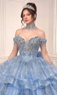 A woman wearing Ladivine’s Quinceanera Dress 15742, a light blue off-the-shoulder beaded gown paired with a lace choker and tiara, stands against a light-paneled background.