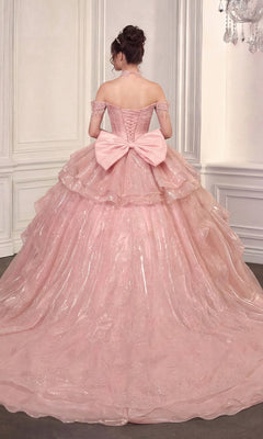 A woman wears the Quinceanera Dress 15742 By Ladivine—an off-the-shoulder pink gown with a detachable skirt and large back bow—while standing in an ornate room with white paneled walls, facing away.