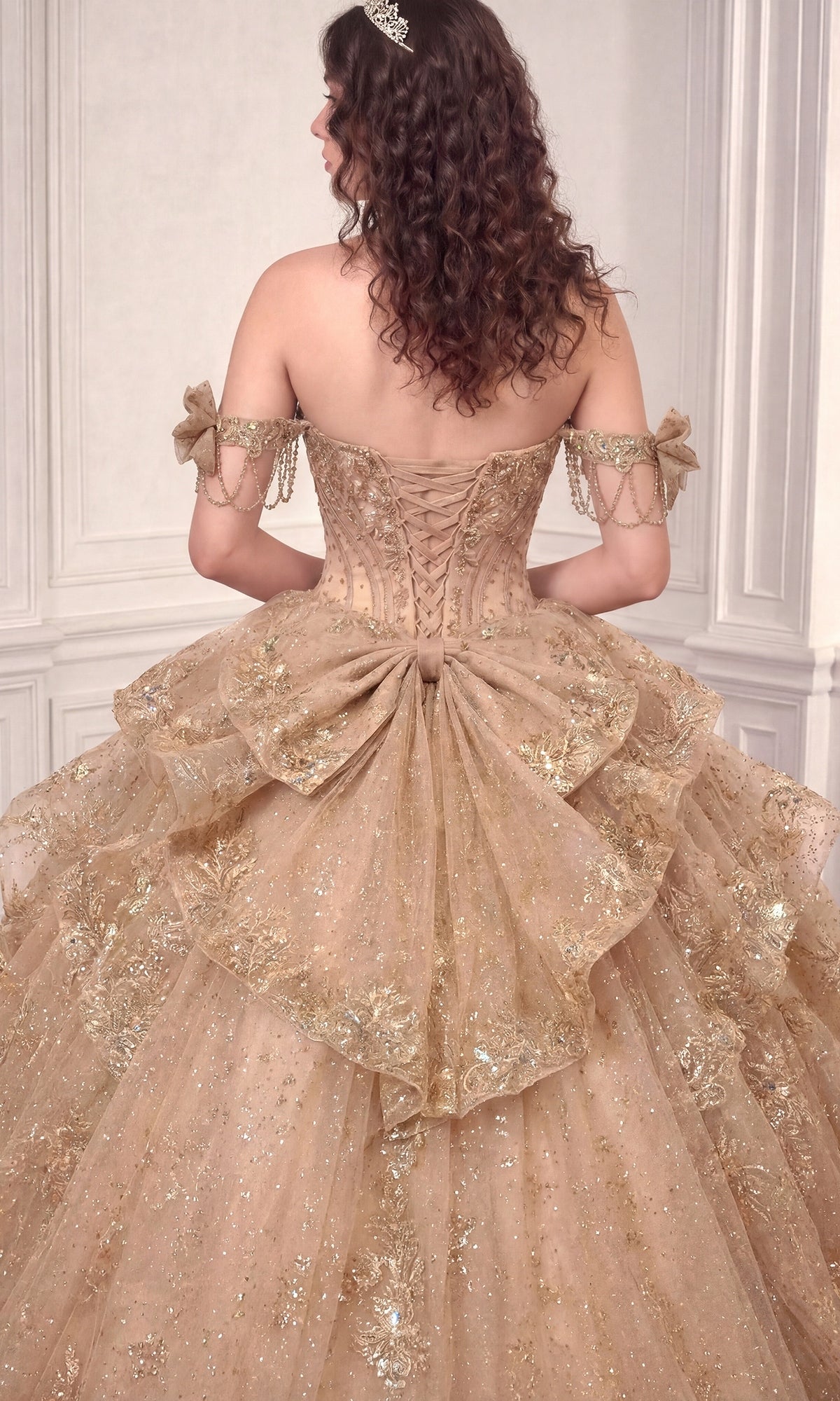 Wearing the Quinceanera Dress 15743 By Ladivine, a person stands facing away in an elegant room. The ornate, off-the-shoulder gown features a lace-up back and glittering beaded accents, with its shimmering gold fabric adding a touch of grandeur to the scene.