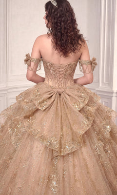 Wearing the Quinceanera Dress 15743 By Ladivine, a person stands facing away in an elegant room. The ornate, off-the-shoulder gown features a lace-up back and glittering beaded accents, with its shimmering gold fabric adding a touch of grandeur to the scene.