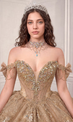 A young woman wearing the Quinceanera Dress 15743 By Ladivine, an ornate gold ballgown adorned with floral embellishments, paired with a jeweled necklace and a silver tiara, stands against a light-colored background.