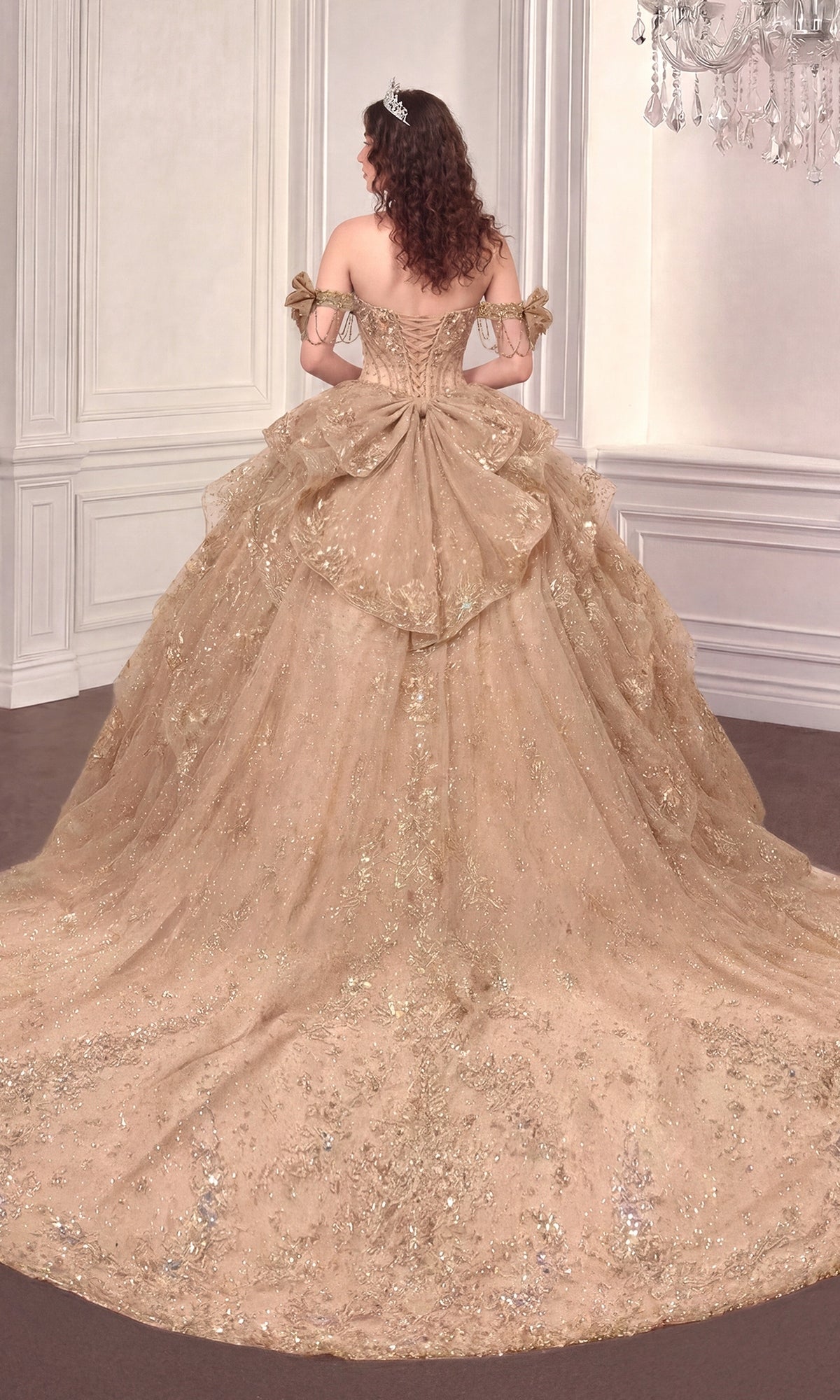 A woman in the Quinceanera Dress 15743 By Ladivine, an off-the-shoulder gold ball gown with a long, layered glittery train, stands in an ornate room with her back to the camera.