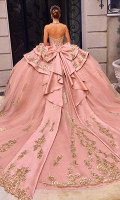 A woman in the Quinceanera Dress 15744 By Ladivine, featuring a pink ballgown silhouette with gold embroidery and a large bow, stands facing away in front of an ornate building entrance.