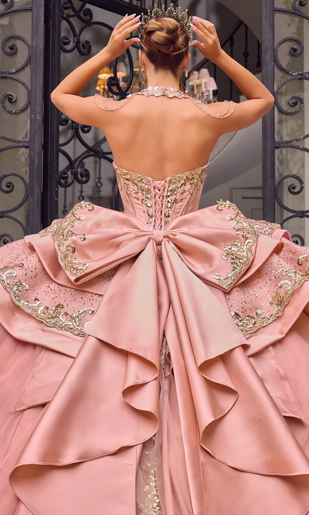 A person stands with their back to the camera, adjusting a jeweled crown while wearing the Quinceanera Dress 15744 By Ladivine—an ornate pink ballgown featuring a large bow and gold embellishments.