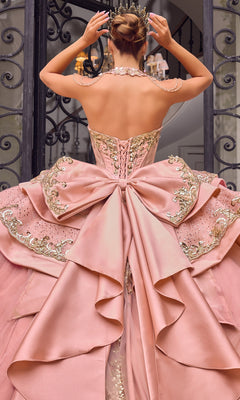 A person stands with their back to the camera, adjusting a jeweled crown while wearing the Quinceanera Dress 15744 By Ladivine—an ornate pink ballgown featuring a large bow and gold embellishments.