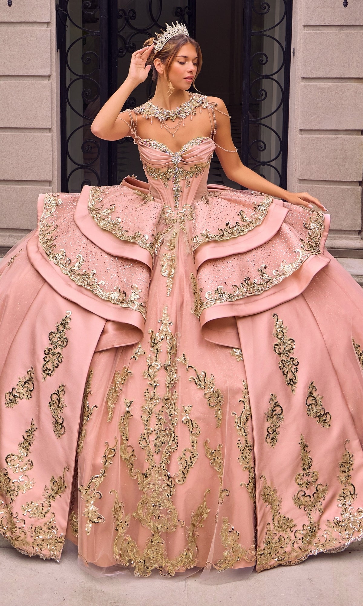A woman wearing the Quinceanera Dress 15744 By Ladivine in rose gold, adorned with intricate gold embroidery and paired with a tiara, stands in front of a stone building with iron doors.