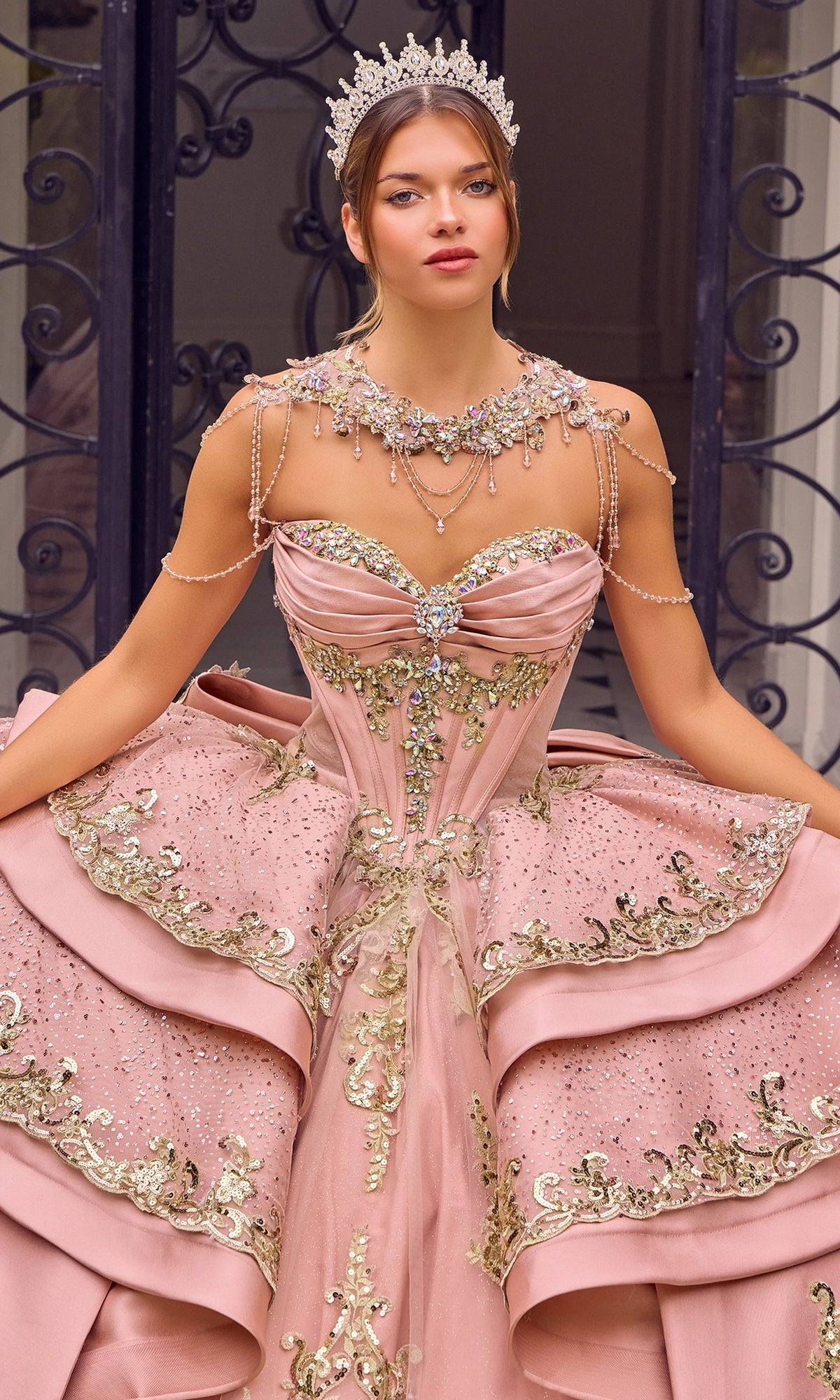 A woman wearing the Quinceanera Dress 15744 By Ladivine—a pink ballgown adorned with gold embroidery and jeweled accents—paired with a sparkling tiara and matching jewelry, stands in front of ornate iron gates.
