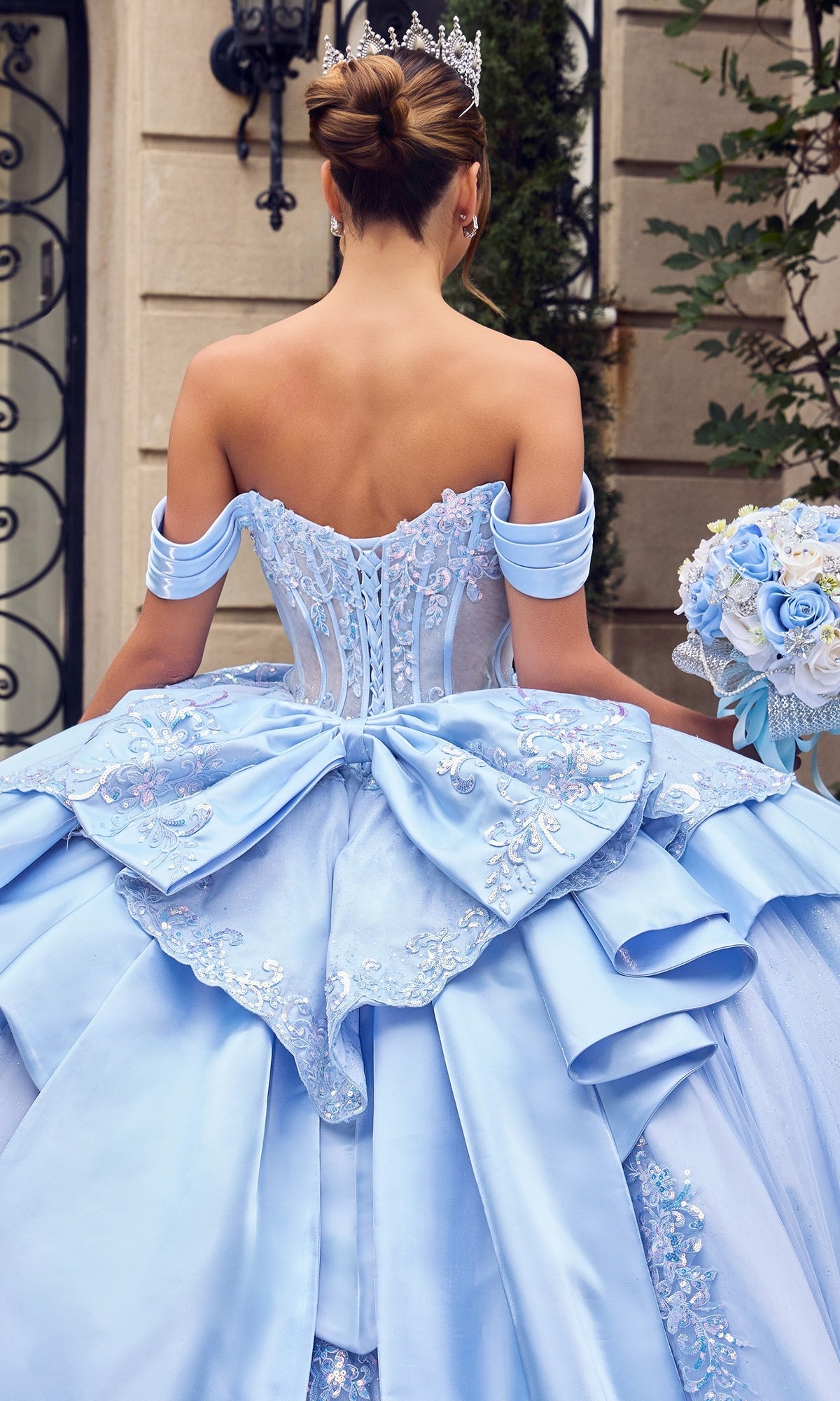 Wearing the Quinceanera Dress 15745 By Ladivine, a woman stands outside holding a pastel bouquet. Her light blue ballgown features a beaded corset bodice and off-shoulder sleeves, while the glitter-tulle skirt shimmers softly in the daylight, adding a magical touch to her elegant look.