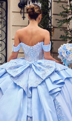 Wearing the Quinceanera Dress 15745 By Ladivine, a woman stands outside holding a pastel bouquet. Her light blue ballgown features a beaded corset bodice and off-shoulder sleeves, while the glitter-tulle skirt shimmers softly in the daylight, adding a magical touch to her elegant look.