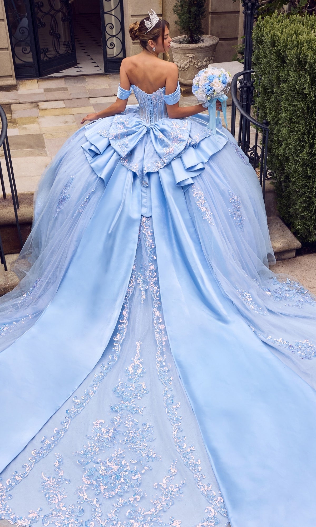 Wearing the Quinceanera Dress 15745 By Ladivine, a woman stands on outdoor steps in a blue ballgown featuring a beaded corset bodice and long embroidered train, holding a bouquet and wearing a tiara.