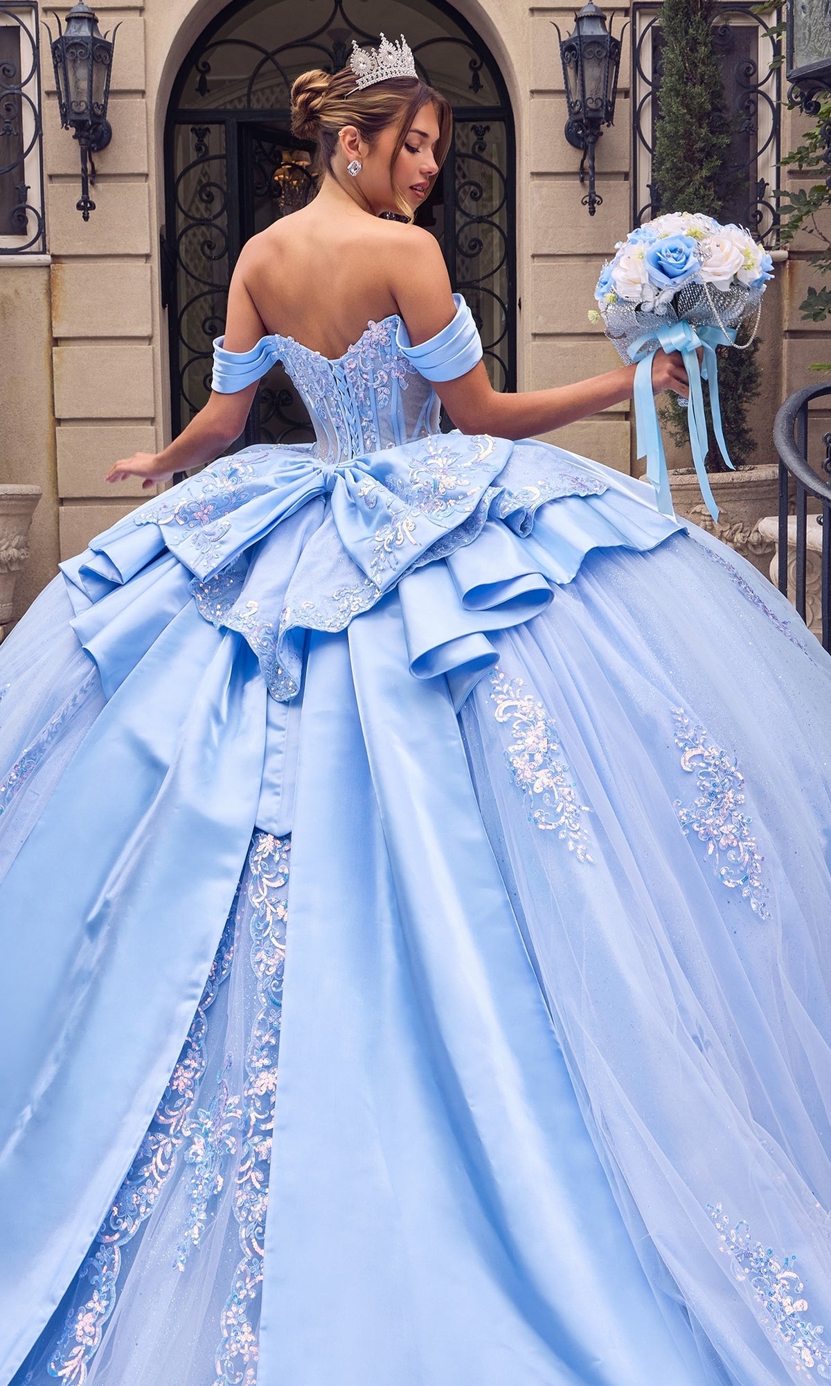 A woman in the Quinceanera Dress 15745 By Ladivine, featuring a blue ballgown silhouette with a beaded corset bodice and embroidered details, stands outside an ornate building holding a bouquet and wearing a crown.