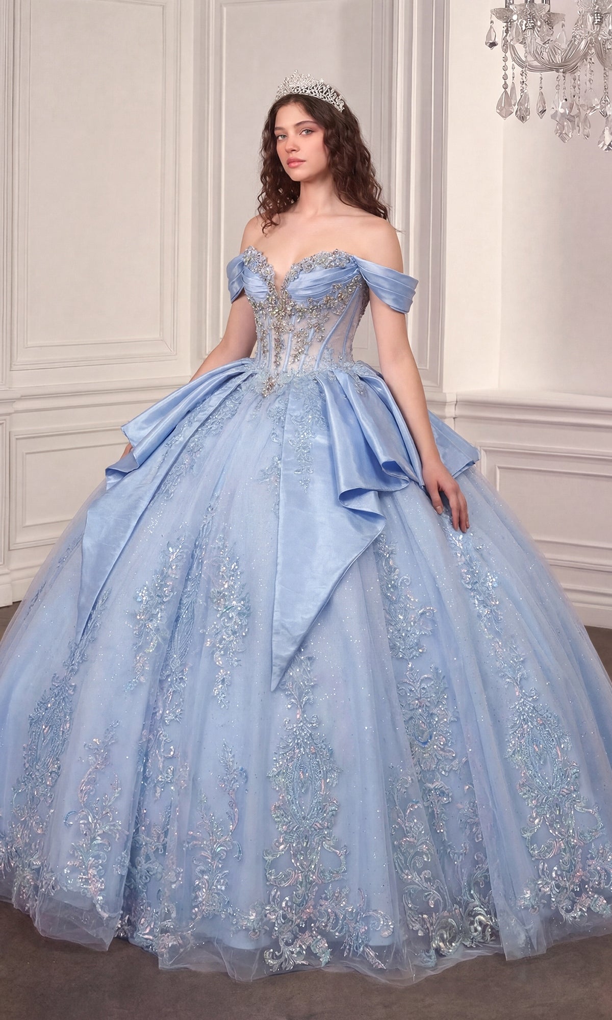 A woman stands in an ornate room with white paneled walls and a chandelier, crowned with a tiara, wearing the Quinceanera Dress 15745 by Ladivine—a sparkling, off-the-shoulder light blue ballgown featuring a beaded corset bodice and a glitter-tulle skirt.