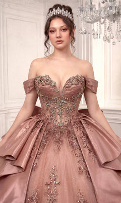 A woman wearing the Quinceanera Dress 15745 By Ladivine, an ornate off-shoulder pink ballgown featuring floral embellishments and a beaded corset bodice, paired with a tiara, stands indoors against white panel walls and beneath a chandelier.