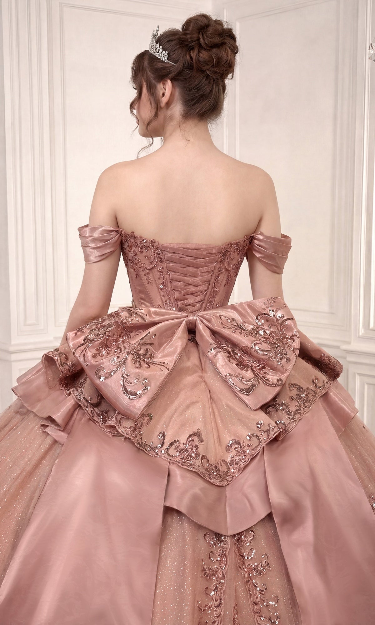 A woman stands facing away in an elegant, white-paneled room, wearing the Quinceanera Dress 15745 By Ladivine—an off-shoulder pink ballgown adorned with a large decorative bow and shimmering sequins. The dress features a beaded corset bodice and a flowing glitter-tulle skirt that enhances the sophisticated scene with sparkling allure.