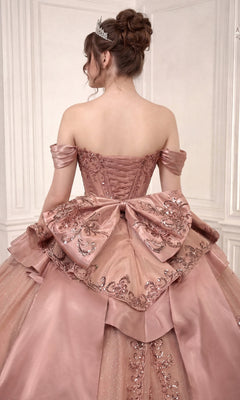 A woman stands facing away in an elegant, white-paneled room, wearing the Quinceanera Dress 15745 By Ladivine—an off-shoulder pink ballgown adorned with a large decorative bow and shimmering sequins. The dress features a beaded corset bodice and a flowing glitter-tulle skirt that enhances the sophisticated scene with sparkling allure.