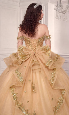 A woman in a gold, off-the-shoulder ballgown adorned with sequins and a large bow stands facing away in an elegant white room, showcasing the stunning silhouette of the Quinceanera Dress 15746 by Ladivine.