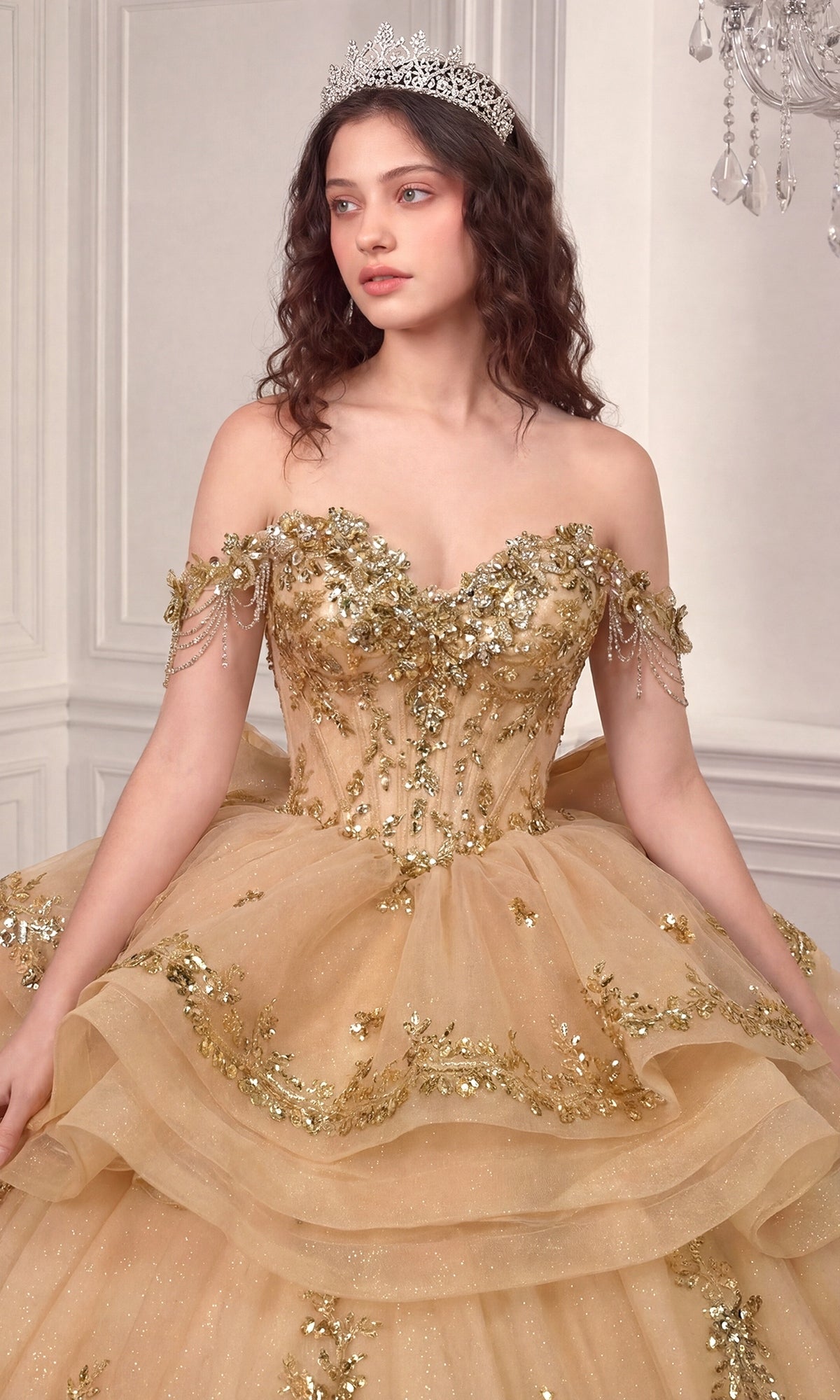 A woman adorned in the Quinceanera Dress 15746 by Ladivine—a lavish gold ballgown with floral embellishments and a tiara—stands gracefully in an elegant room. The off-the-shoulder design of this regal Ladivine dress beautifully enhances her graceful presence.