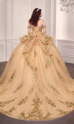 A woman stands gracefully in an elegant, light-colored room wearing the Quinceanera Dress 15746 By Ladivine—an off-the-shoulder ball gown featuring gold embellishments and a large bow on the back.