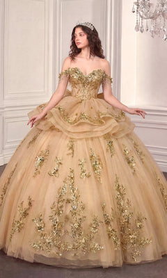 A woman wearing the Quinceanera Dress 15746 By Ladivine in Champagne Gold—with its off-the-shoulder silhouette and floral embellishments—stands in an elegant room featuring white paneled walls and a chandelier, radiating regal charm.