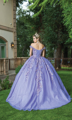 A woman in the Quinceanera Dress 1574 by Dancing Queen, featuring a sweetheart neckline and floral embroidery, stands on grass facing away near a building and tall trees.