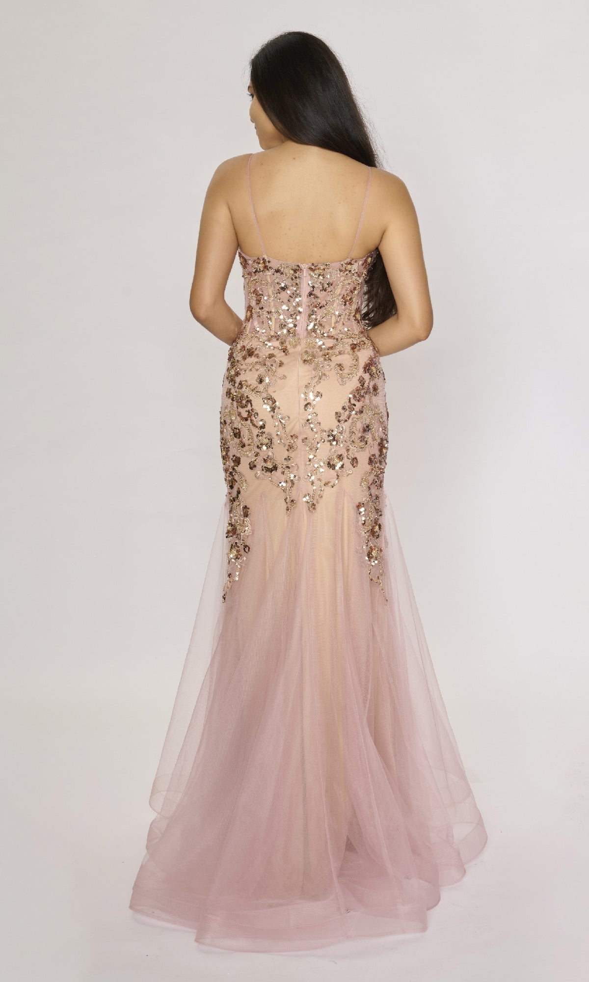 A woman stands with her back to the camera, wearing the Nina Canacci 1576 long formal dress—a sleeveless light pink mermaid gown with floral sequin embellishments and flowing tulle.