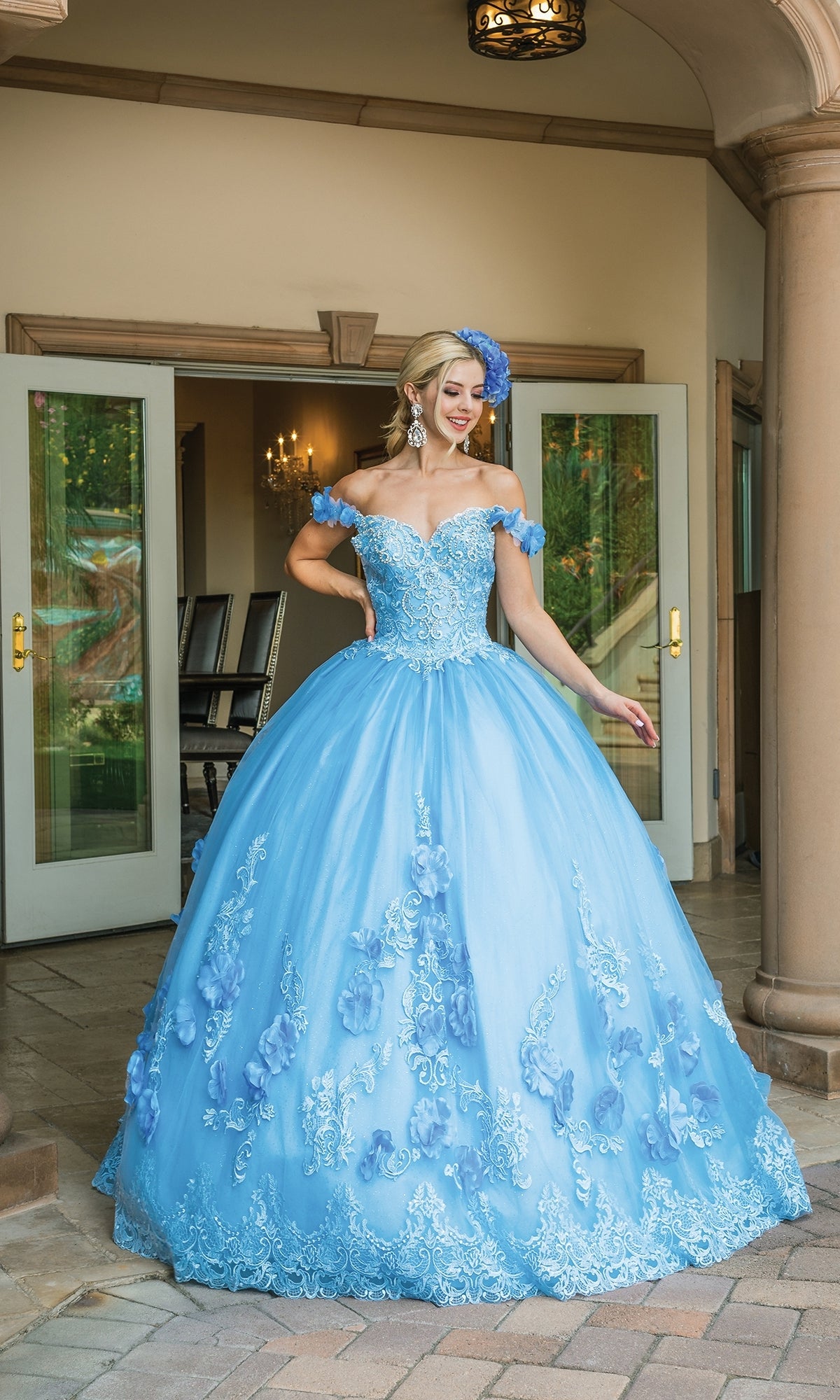 A woman smiles outside in front of open doors, wearing the Quinceanera Dress 1582 by Dancing Queen—a light blue, a-line ball gown with floral details, off-the-shoulder straps, and a matching headpiece.