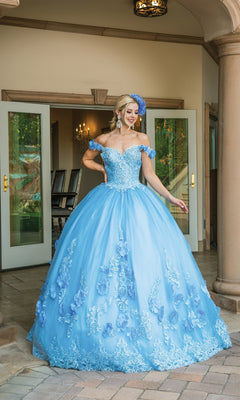 A woman smiles outside in front of open doors, wearing the Quinceanera Dress 1582 by Dancing Queen—a light blue, a-line ball gown with floral details, off-the-shoulder straps, and a matching headpiece.