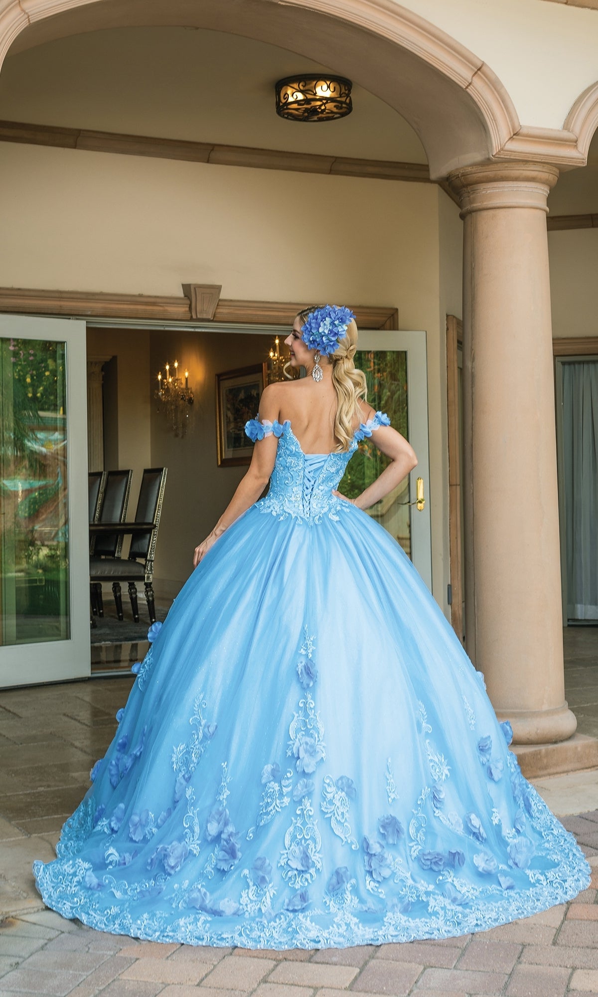 A woman stands outdoors, facing away, wearing the Quinceanera Dress 1582 by Dancing Queen—an a-line ball gown with floral appliqués, off-the-shoulder straps, and a matching blue headpiece.