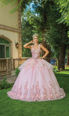 A woman poses on grass in an outdoor garden, wearing Quinceanera Dress 1583 by Dancing Queen—a pink ball gown adorned with floral embroidery and a tiara, with one hand on her hip.