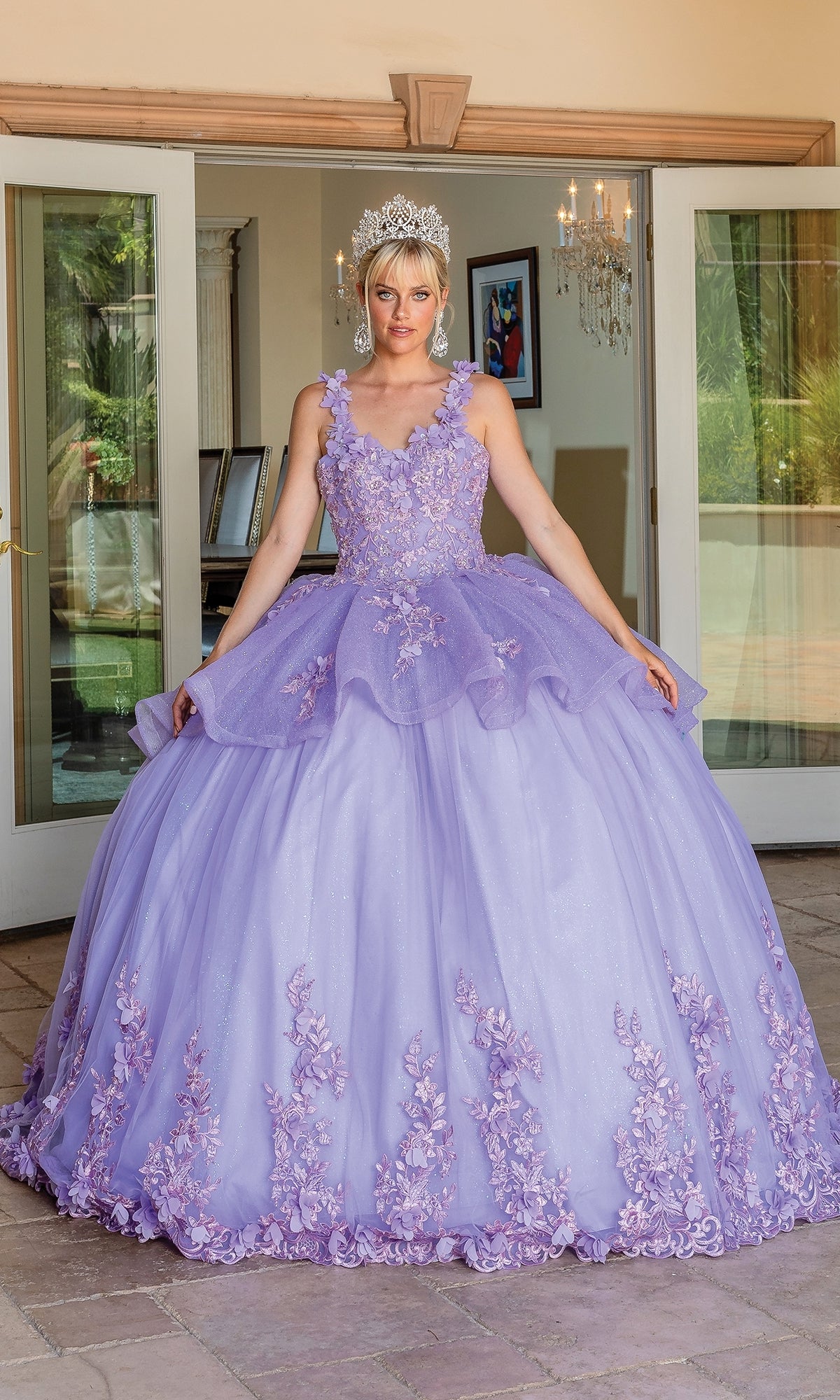 A woman wearing the Quinceanera Dress 1583 by Dancing Queen, a lavender ball gown with floral embellishments and a tiara, stands in front of open glass doors.