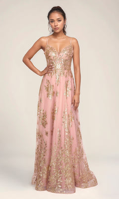A woman models the Nina Canacci 1589, a pink floor-length formal gown with an embellished bodice, thin straps, gold floral details, and an elegant a-line skirt, set against a plain light background.