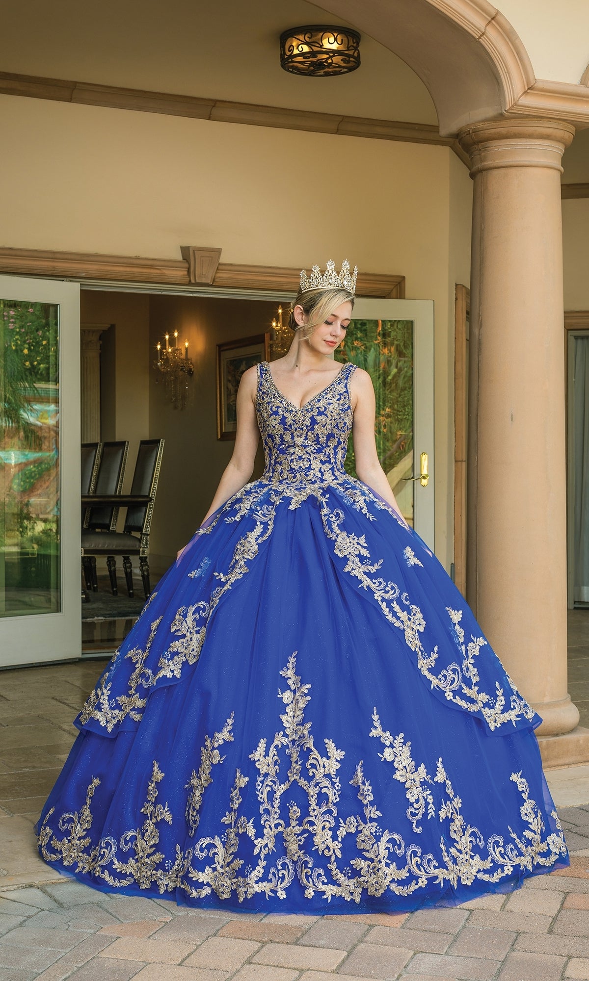 A woman stands outside near open double doors wearing the Quinceanera Dress 1594 by Dancing Queen, a blue ball gown featuring gold embroidery and topped with a matching gold crown.