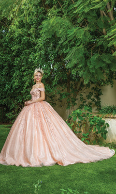 A woman smiles at the camera while standing on grass near tall green trees, wearing a tiara and the Quinceanera Dress 1595 by Dancing Queen—a pink, off-the-shoulder ball gown with a sweetheart neckline.