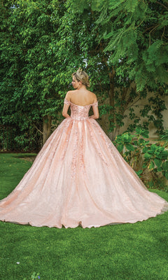 A woman wearing the Quinceanera Dress 1595 by Dancing Queen, a sparkly pink a-line gown with off-the-shoulder straps, stands on grass with her back to the camera, surrounded by lush greenery and trees.