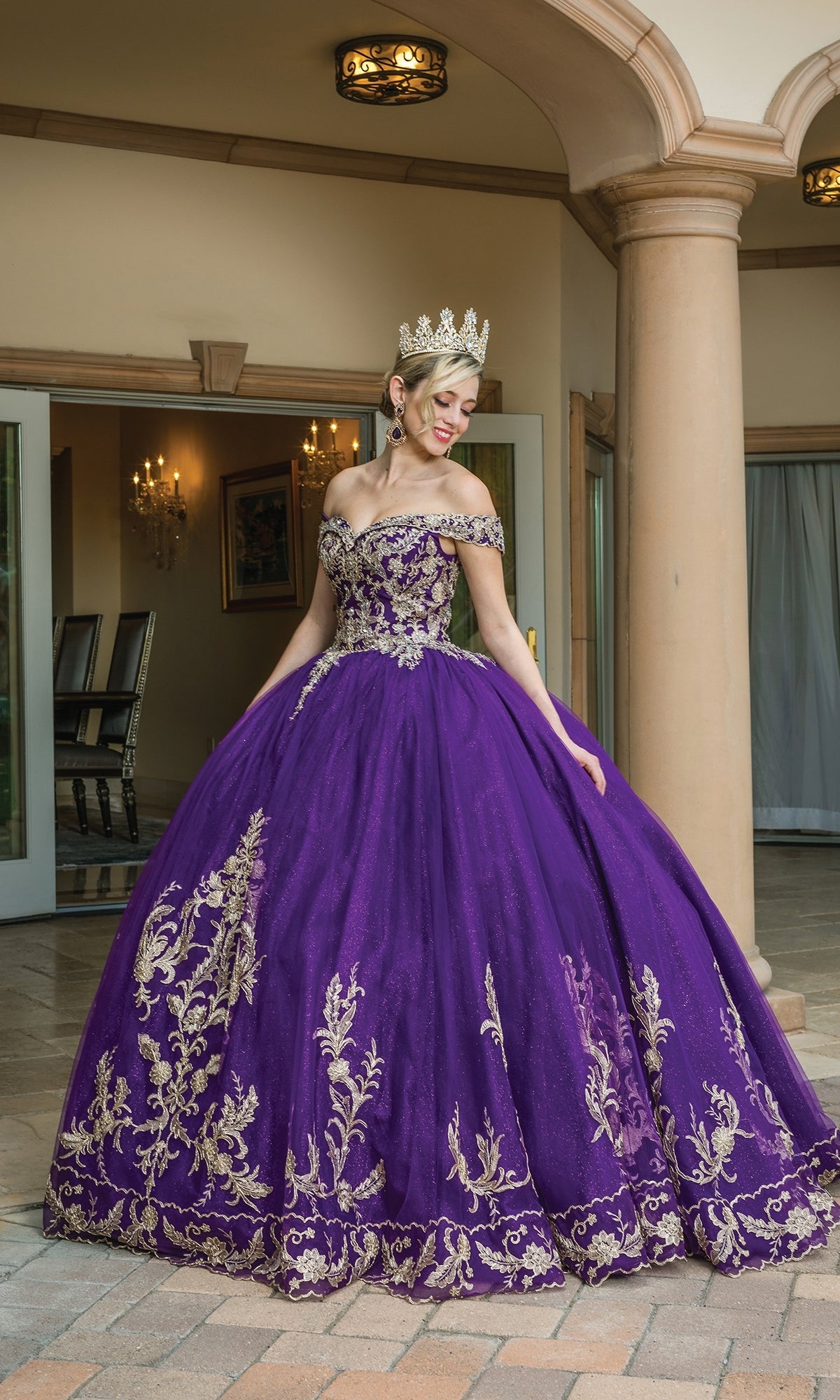 A woman in the Quinceanera Dress 1596 by Dancing Queen, a purple a-line ball gown with gold embroidery and off-the-shoulder straps, stands outside a columned building wearing a jeweled crown.