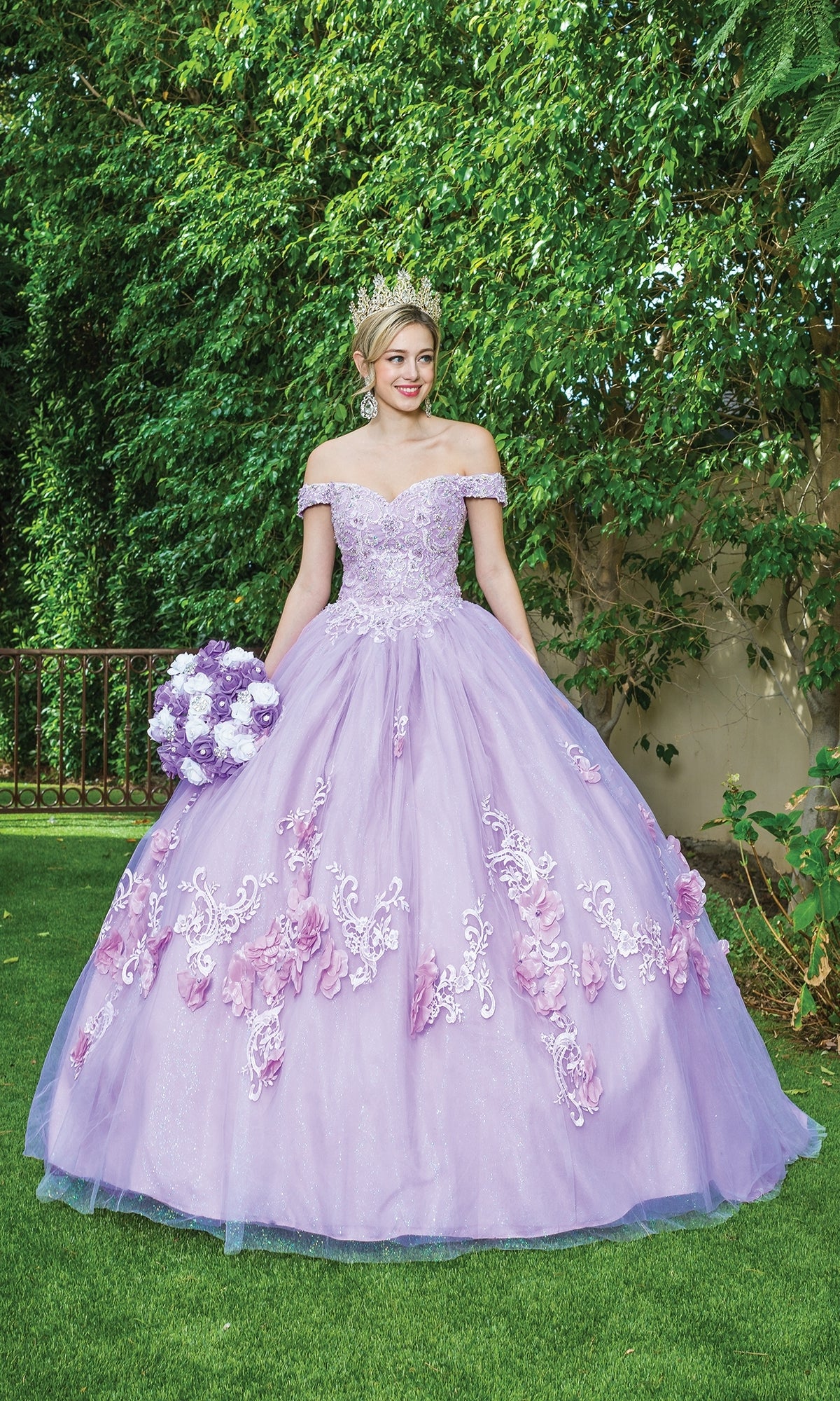 Young woman wearing the Quinceanera Dress 1598 By Dancing Queen—an off-shoulder lavender ball gown with floral accents and a tiara—stands on grass holding a bouquet, with greenery and a fence in the background.