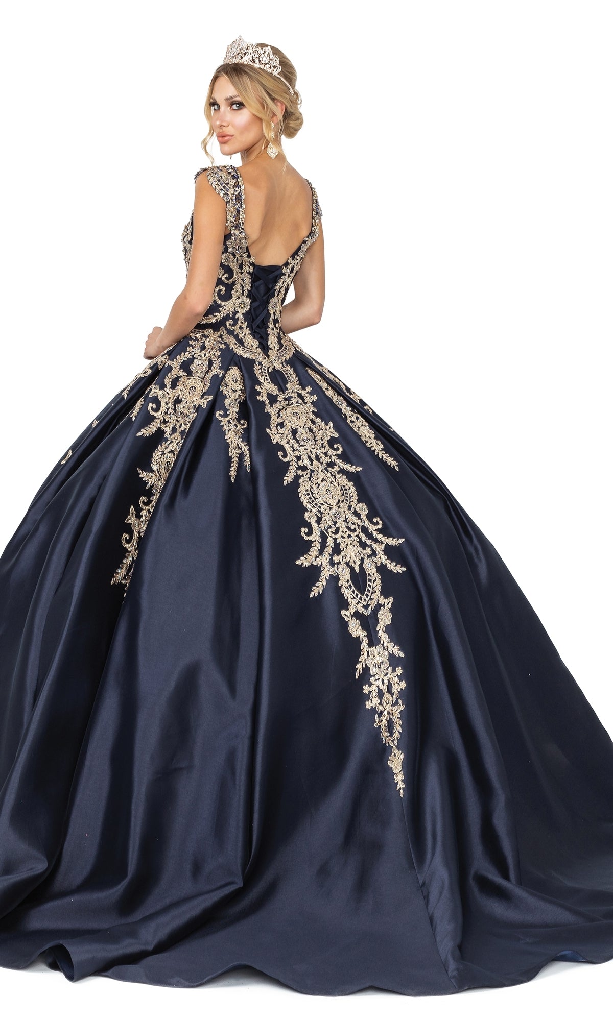 A woman in Quinceanera Dress 1606 by Dancing Queen, featuring navy blue fabric, gold embroidery, cap sleeves, and a tiara, poses against a white background.
