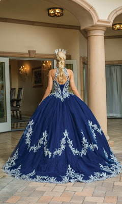 A person wearing the High-Neck Navy Quinceanera Dress 1607, a floor-length gown with silver lace embroidery and a crown, stands facing away in an elegant courtyard.