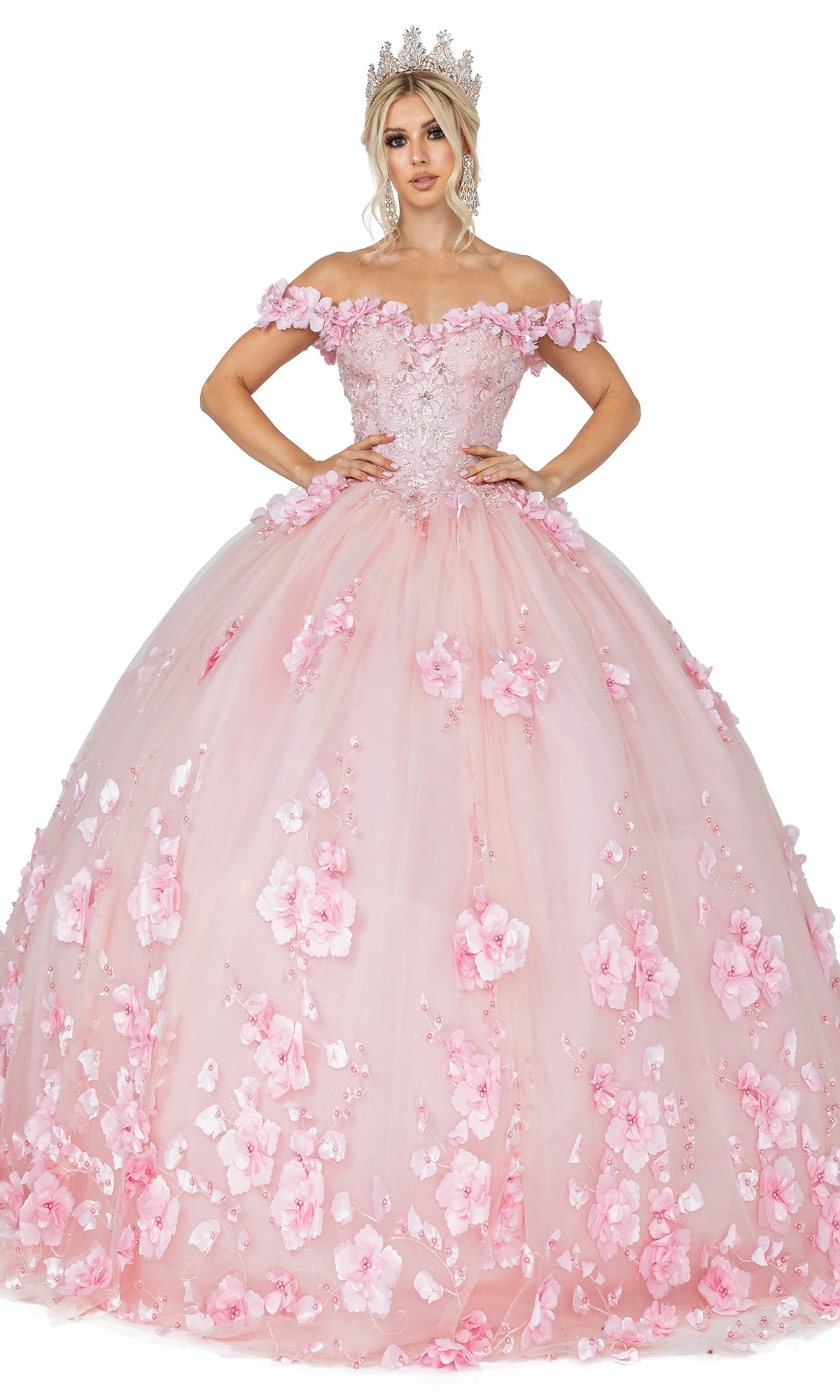A woman poses with hands on hips in the Quinceanera Dress 1613 by Dancing Queen, a pink off-the-shoulder ball gown featuring floral appliqués and a tiara, set against a white background.
