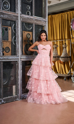 A woman stands indoors before metal speakers, wearing the Tiffany 16144 long prom dress, featuring a pink sweetheart neckline, thin straps, front cutout, and ruffled A-line skirt. Chandeliers and yellow curtains decorate the background.