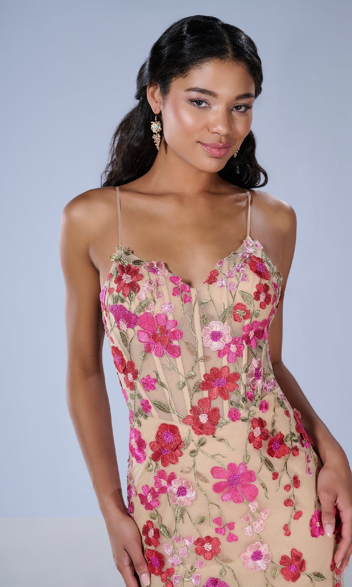 A woman wears the Tiffany 16148 Long Prom Dress, a sleeveless beige gown with pink and red floral embroidery and delicate lace applique, standing against a plain light blue background.