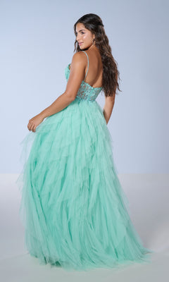 A woman stands slightly turned in the Tiffany 16149 long prom dress, featuring a light turquoise tulle skirt with cascading ruffles and a fitted, embellished bodice, against a plain background.