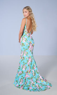 A woman poses with her back to the camera against a plain light blue background, wearing the Tiffany 16155 long prom dress—a turquoise, floral-patterned, backless gown featuring a thigh-high slit.