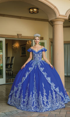 A woman in the Off-the-Shoulder Quinceanera Dress 1615, featuring blue and silver lace embroidery and a crown, stands before an open doorway with columns and gleaming chandeliers inside.