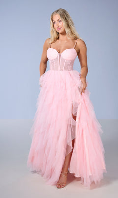 A woman wears the Tiffany 16161 long prom dress, featuring light pink tulle, spaghetti straps, a fitted corset bodice, and a high slit, standing against a plain light blue background.